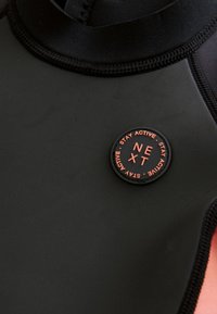 Black neoprene wetsuit material featuring a round logo patch in orange with text "STAY ACTIVE" and "NEXT." Smooth texture with fitted design.