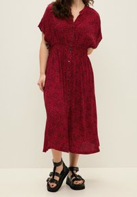 Rotes Maxikleid mit kurzen Ärmeln, Knopfleiste vorn, geraffter Taille und einem dezenten Muster. Kombiniert mit schwarzen Riemchensandalen.