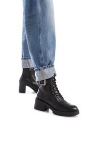 Botas de cuero negras hasta el tobillo con un tacón grueso y un diseño de cordones. Acabado texturizado con costuras prominentes y un detalle de cremallera lateral.