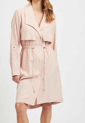 Kobieta w jasnoróżowym płaszczu typu trench do kolan z długimi rękawami i talią ze sznurkiem na gładkim tle.