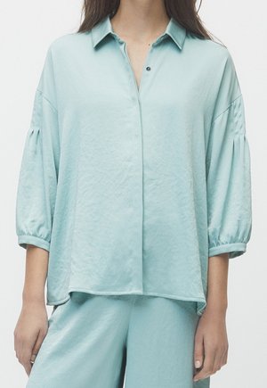 Vrouw draagt een losse, lichtblauwe satijnen blouse met driekwart pofmouwen en bijpassende wijde broek tegen een effen achtergrond.
