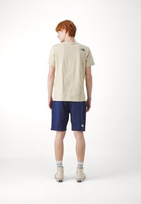 T-shirt in cotone beige chiaro con maniche corte e logo nero sul retro, abbinato a pantaloni corti blu scuro e scarpe in tela beige.
