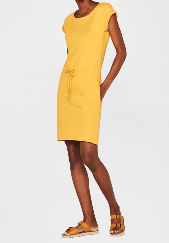 Femme portant une robe jaune au genou avec une ceinture à la taille, des manches courtes et des sandales jaunes à boucle, debout avec une main dans la poche.