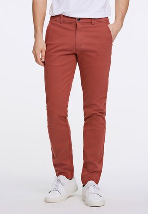 Mann trägt rostfarbene Slim-Fit-Hosen und weiße Sneaker, steht mit einer Hand in der Tasche vor einfachem Hintergrund.