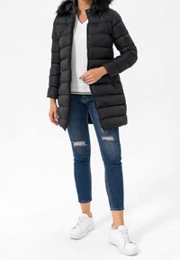 Sort pufferjakke med hætte i faux fur, quiltet design og lynlås. Parret med en hvid kabelstrikket sweater, slidte jeans og hvide sneakers.