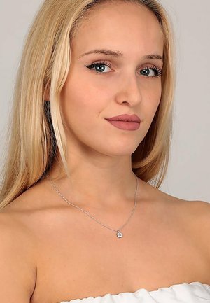 Silberne Kettenhalskette mit einem kleinen Herzanhänger, auf einer nackten Schulter präsentiert. Der Hautton ist hell, das Haar lang und blond.