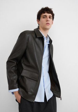 Junger Mann trägt eine schwarze Lederjacke über einem hellblauen Hemd, mit den Händen in den Taschen, steht vor einer weißen Wand.