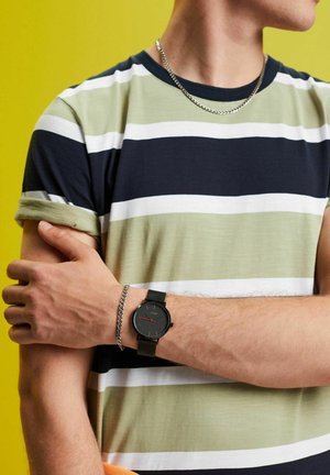 Schwarz Uhr mit roten Akzenten am Handgelenk, getragen über einem grün-blau gestreiften T-Shirt, mit einer silbernen Kettenhalskette, vor einem gelben Hintergrund.