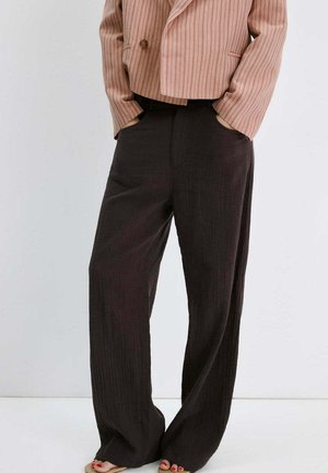 Mujer con blazer corto de rayas rosas y pantalones negros sueltos de talle alto, de pie con las manos en los bolsillos, visible desde los hombros hacia abajo.