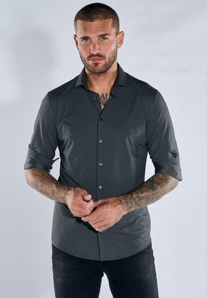 Graues Langarmshirt aus glattem Stoff, mit Knopfleiste, aufgerollten Ärmeln und abgerundetem Saum. Schwarze Hose sichtbar.