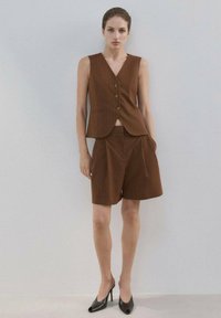 Mujer de pie contra una pared lisa, vestida con chaleco marrón de rayas finas sin mangas y pantalones cortos sueltos a la altura de la rodilla a juego, con tacones altos negros.