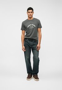 Γκρι βαμβακερό t-shirt με λογότυπο "TOMMY JEANS", συνδυασμένο με σκούρα μπλε τζιν και καφέ αθλητικά παπούτσια. Το μοντέλο στέκεται στραμμένο μπροστά.