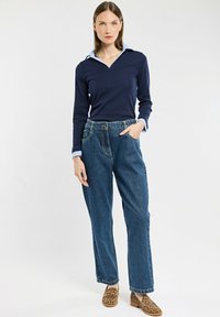 Donna in piedi che indossa un top a maniche lunghe a strati blu navy, jeans a vita alta blu e mocassini con stampa leopardo, su uno sfondo bianco semplice.