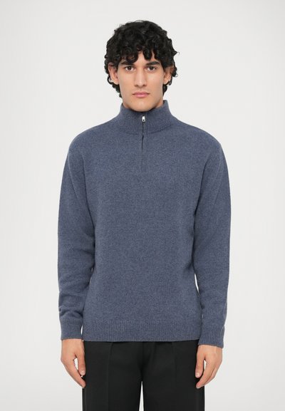 Mann mit lockigem Haar, der einen blauen Strickpulli mit halbem Reißverschluss und schwarze Hosen trägt, steht vor einem schlichten hellen Hintergrund.