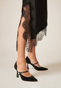 Svart satinklänning med spetskant, med invecklade mönster. Kompletteras med svarta högklackade skor dekorerade med glittrande detaljer och en rem.