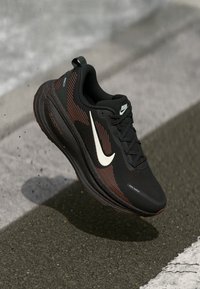 Black and brown Nike running shoe with white swoosh logo, black laces, and cushioned sole on textured pavement.