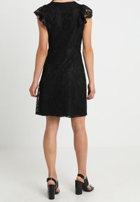 Svart spetsklänning med korta volangärmar, åtsittande midja och utställd nederkant. Bärs med svarta högklackade sandaler. Texturen synlig.