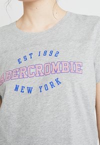 Graues Baumwoll-T-Shirt mit "ABERCROMBIE" in blauen und pinkfarbenen Buchstaben, darüber in einer geschwungenen Schriftart bestickt "EST 1892" und "NEW YORK".