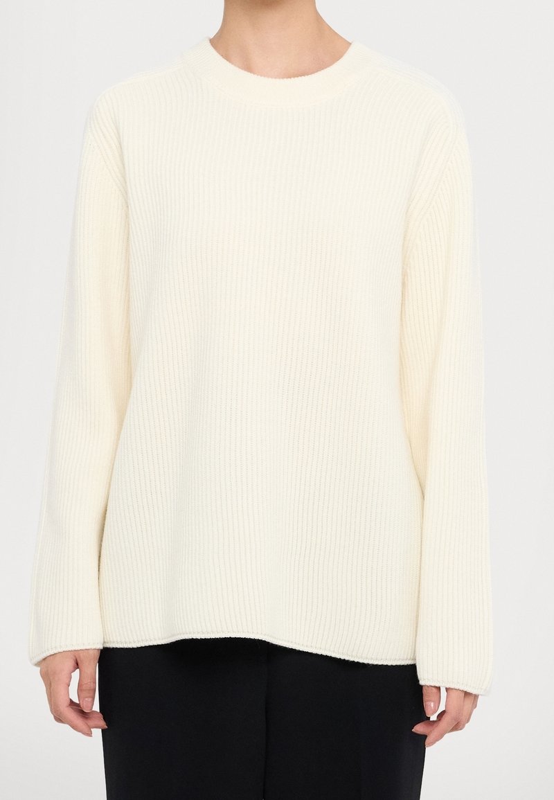 Pull crème côtelé avec encolure ronde, coupe ample et manches longues. Texture douce, avec un ourlet droit et de petites fentes latérales.