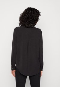 Blusa negra de manga larga con dobladillo curvado, puños abotonados, detalle de pliegue en la parte posterior y un tejido suave y texturizado.