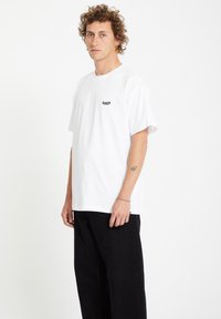 Weißes Baumwoll-T-Shirt mit kurzen Ärmeln, ausgestattet mit einem kleinen schwarzen Logo auf der Brust und einem lockeren Schnitt. Kombiniert mit schwarzen Hosen.