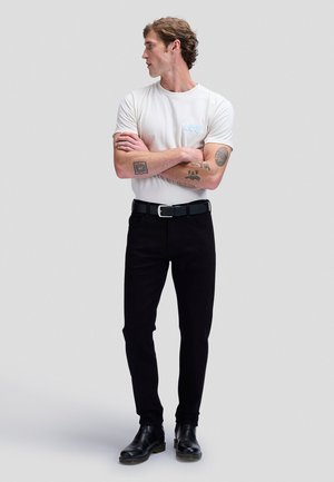 Ung mand med tatoveringer iført en hvid Lee t-shirt, sorte bukser, sort bælte og sorte støvler, står med armene over kors og ser til venstre.