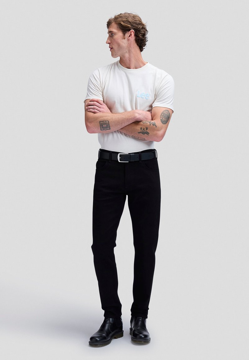Young man with tattoos wearing a white Lee t-shirt, black pants, black belt, and black boots, standing with arms crossed looking left.