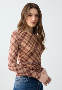 Langärmelige top van transparante beige stof met een bruin ruitpatroon. Heeft een boothals en geplooide details bij de taille. Gepaard met blauwe jeans.