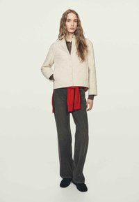 Prošívaná krémová bunda s knoflíkovým zapínáním, doplněná červeným pásem, hnědou košilí a tmavými širokými kalhotami. Texturovaná látka, volný střih.