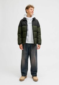 Chaqueta acolchada en negro y verde oliva con diseño acolchado, llevada sobre un sudadera gris. Complementada con unos vaqueros holgados azules y zapatos beige.