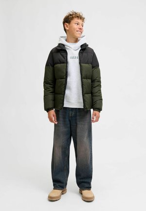 Pufferjack in zwart en olijfgroen met een doorgestikt ontwerp, gedragen over een grijze hoodie. Compleet met losse blauwe jeans en beige schoenen.