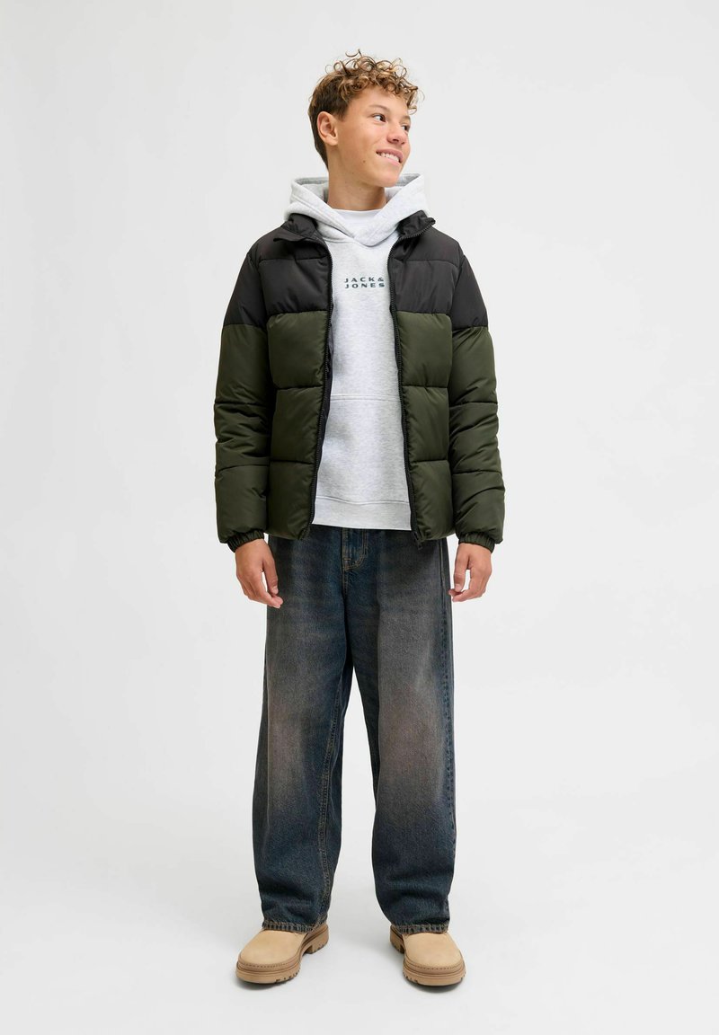 Chaqueta acolchada en negro y verde oliva con diseño acolchado, llevada sobre un sudadera gris. Complementada con unos vaqueros holgados azules y zapatos beige.