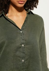 Camicia verde oliva in lino leggero con colletto morbido, texture sottile e bottoni in tonalità argento sulla parte anteriore.
