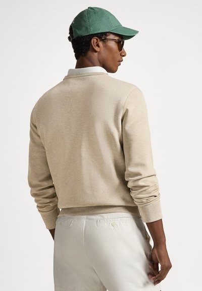 Μπεζ βαμβακερό φούτερ με μακριά μανίκια, ριμπ μανσέτες και άνετη εφαρμογή, σε συνδυασμό με σορτς σε ανοιχτό χρώμα και πράσινο καπέλο.