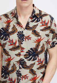 Camisa de manga corta en beige claro con un patrón tropical de hojas verde oscuro y rojas. Cuenta con un cuello de botón y botones blancos.