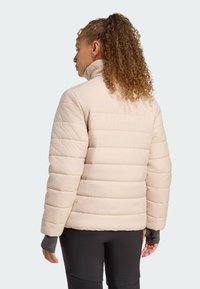 En ung person med krøllet hår har en lys beige oppustet jakke på og står med ryggen til. Baggrunden er ensfarvet og lys.