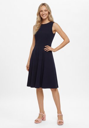 Vrouw glimlachend, gekleed in een mouwloze marineblauwe jurk en roze hoge hak sandalen, staand met één hand op de heup tegen een witte achtergrond.