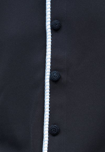 Melnas auduma krekls ar centrālu pogu rindu, kurā ir apaļas tumši zila krāsas pogas, un balt-zilu šuvju detaļas gar malu.