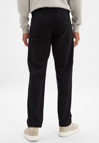 Pantalon en coton noir à coupe droite, avec deux poches arrière et une taille sans couture, associé à des baskets de couleur claire.