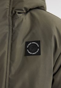 Jachetă de culoare verde măslin cu textură matlasată, având un patch logo pătrat negru cu text alb. Include detalii cu cusături vizibile.