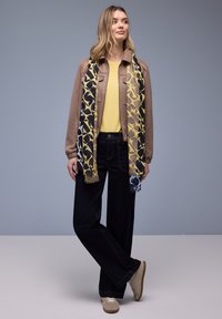 Femme debout, jambes croisées, portant une veste beige, un haut jaune, une écharpe à motifs, un pantalon large sombre et des baskets beiges sur fond gris.