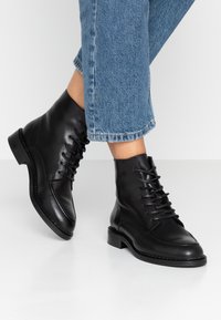 Botines de cuero negro hasta el tobillo con punta redonda, cordones en la parte delantera y superficie texturizada. Presentan un tacón bajo apilado y costuras decorativas.