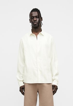 Homme avec des cheveux tressés portant une chemise blanche ample à manches longues et un pantalon beige, debout devant un fond blanc uni.