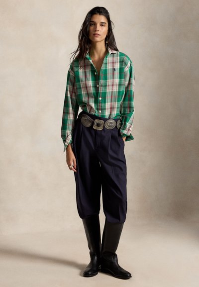 Femme portant une chemise à carreaux verte boutonnée, un pantalon bleu marine avec une large ceinture décorative et des bottes noires montant jusqu'aux genoux, debout devant un fond neutre.