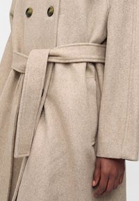 Gros plan d'un manteau en laine beige à boutons croisés et ceinture nouée, montrant la main d'une personne posée sur le côté.