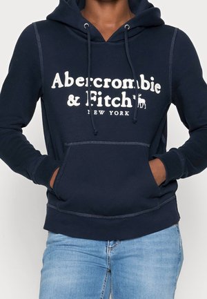 Osoba ubrana w granatową bluzę z kapturem Abercrombie & Fitch z kieszenią kangura i jasnoniebieskie dżinsy, ręce częściowo w kieszeniach.
