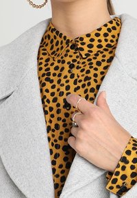 Leopardikuviollinen paita, jossa mustia pilkkuja sinapinvärisellä taustalla, napitettu ylös, päällä vaaleanharmaa takki. Klassinen kaulus.