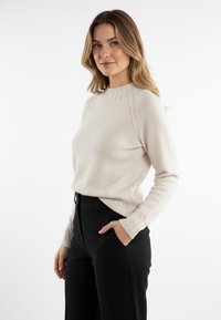 Jasnobeżowy sweter z dzianiny o okrągłym dekolcie i warkoczowym wzorze na ramionach oraz mankietach, zestawiony z czarnymi, dopasowanymi spodniami.