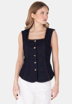 Donna che indossa un gilet senza maniche in denim scuro con bottoni dorati e pantaloni bianchi, guarda di lato su sfondo uniforme.