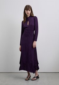 Vestido morado de manga larga con un cuello alto y un recorte en forma de ojo de cerradura, con una textura ondulada y un dobladillo fluido. Modelo con tacones negros.